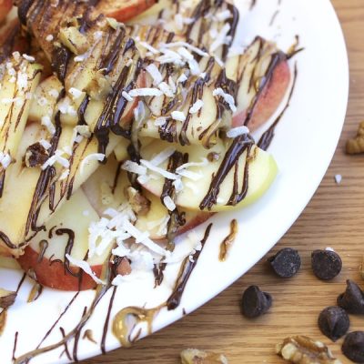 Apple slices topped with drizzles of peanut butter, chocolate topped with nuts and shredded coconut on a plate with chocolate chips and walnuts on the table next to it.