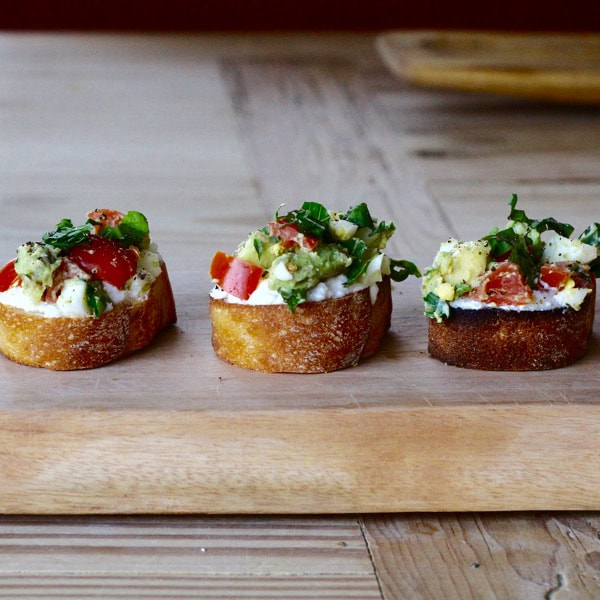 Three sliced whole wheat baguette bread topped with ricotta cheese and fresh vegetables.