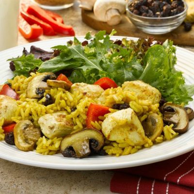 A plate of curried chicken with mushrooms, bell peppers, raisins and rice with a side salad