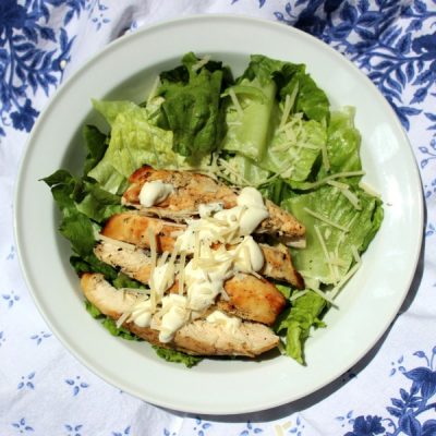 Caesar salad with grilled chicken in a bowl on a white and blue tablecloth.