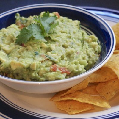 Guacamole garnished with leaves of cilantro in a bowl, on a plate with corn chips.
