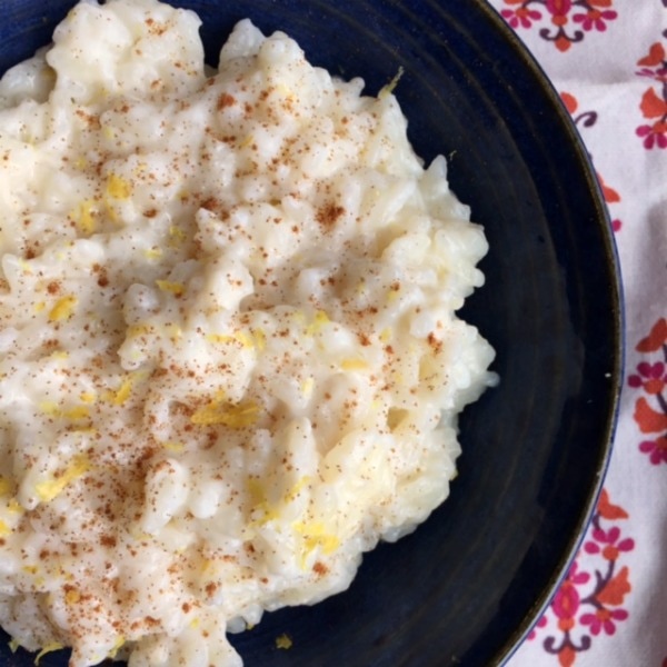 Bowl of homemade rice pudding topped with lemon zest and cinnamon in a dark bowl on a white, red, and orange patterned tablecloth.