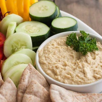 Bowl of hummus with sliced cucumbers and pita bread on the side