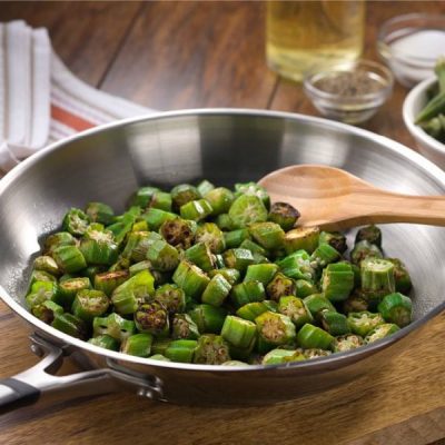 Lightly fried okra in a stainless steel skillet with wooden spoon resting on a wooden cutting board with ingredients and a cloth napkin in the background.