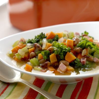 Bowl of mixed vegetable and turkey ham soup topped with fresh parsley on a brightly colored cloth with a spoon on the side.