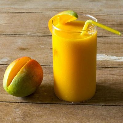 Cup of mango banana smoothie with a straw and a mango wedge on the rim. Mango with slice cut out on the side on a wooden table.