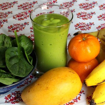 Mango green smoothie in class cup on patterned table cloth surrounded by a mango, oranges, banana, and plate of spinach.