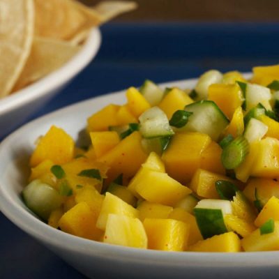 Dish of mango salsa on a blue table cloth with a dish of corn chips in the background.