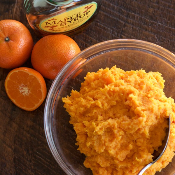 Glass bowl of maple sweet potatoes with a fork. Oranges and bottle of maple syrup on the side all on a wooden table.