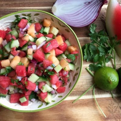 Decorative serving bowl of mixed melon salad topped with chopped cilantro with red onion, watermelon, cilantro, and a lime on the side.