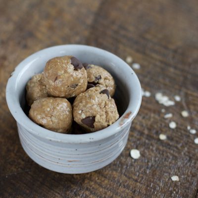Small bowl holding oat balls with chocolate chips in them on a wooden table with a few oatmeal flakes on the side.