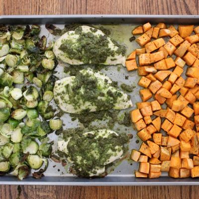 A sheet pan divided into thirds filled with roasted Brussels sprouts on the left, chicken breast topped with pesto in the middle, and roasted sweet potatoes on the right.