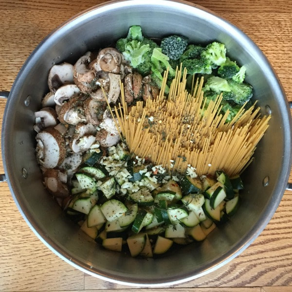 Cooking pot on a wooden table filled with raw spaghetti, broccoli, mushrooms, and zucchini topped with seasoning before cooking.