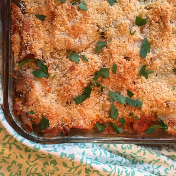 Closeup of a corner of a glass baking dish with chicken parmesan over pasta topped with cheese and chopped parsley on a floral patterned tablecloth.