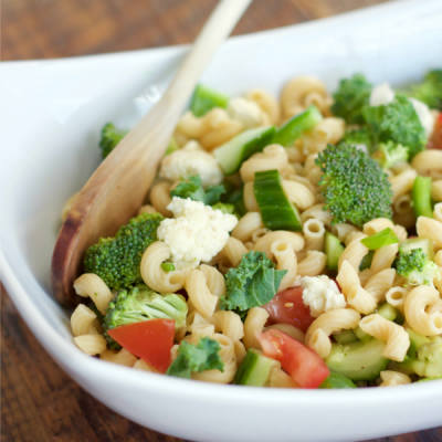 Serving bowl holding pasta salad with mixed vegetables and a mixing spoon on a wooden table.