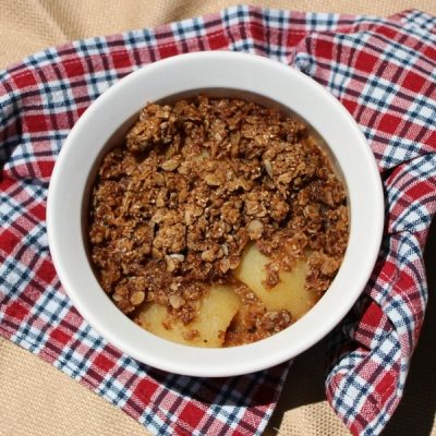 Baking bowl filled with peach cobbler topped with granola on a plaid table cloth.