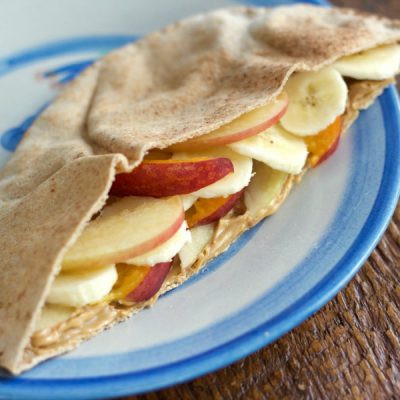 A pita pocket filled with thin slices of apple and banana with peanut butter on a blue plate sitting on a wooden table.