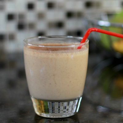 Glass holding a peanut butter banana smoothie with a red straw on a kitchen counter and mosaic backsplash.