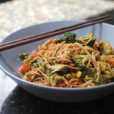 Bowl of noodles and mixed vegetables with peanuts. Two dark wooden chopsticks rest across the bowl.