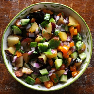 Decorative serving bowl of plum cucumber salad topped with chopped cilantro on a rustic wooden table.