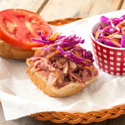 An open slider bun with pulled pork topped with cabbage slaw on one side and a large slice of tomato on the other have of the bun next to a small serving dish filled with cabbage slaw.