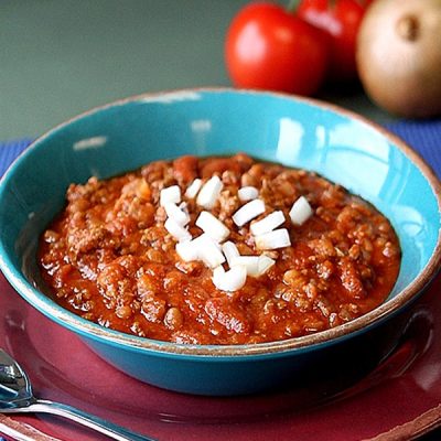 Bowl of chili topped with onion with whole tomatoes and onion in the background.