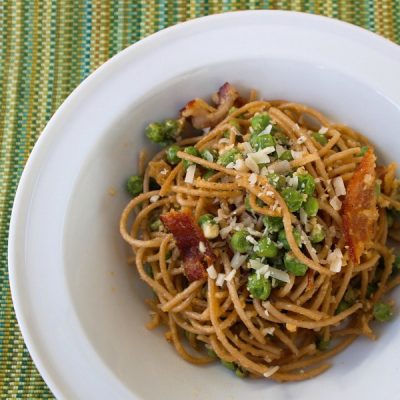 Bowl of whole wheat spaghetti mixed with peas and turkey bacon covered in shredded parmesan cheese.