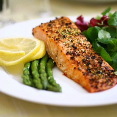 Piece of baked salmon topped with pepper and other spices on a plate with sliced lemon, cooked asparagus and a salad.