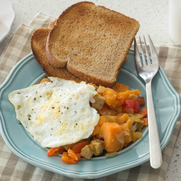 Plate of hash with chopped sweet potatoes, onion, and peppers next to a sunny side down eggs and two slices of wheat toast.