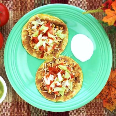 Two small corn tortillas covered with black beans, shredded cheese, and taco sauce with a dollop of sour cream on the side.