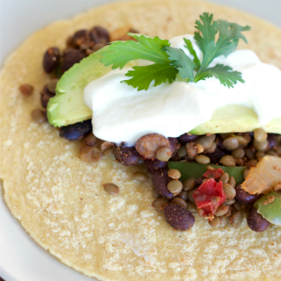 Flour tortilla lying flat covered with cooked lentils, black beans, taco seasoning, topped with sour cream and avocado slices.
