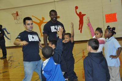 Two male young adults smiling engaging a group of elementary school students in a gym.