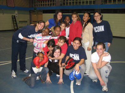Group of young women and girls smiling standing in a gym with boxing equipment.