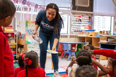 UConn Student hold plate of cut fruit out to group of pre-k students sitting on a classroom carpet.